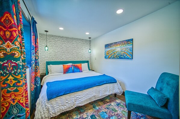 Un dormitorio con una cama, un sillón color turquesa, un tapete colorido, una pared con un diseño de texto y un cuadro.