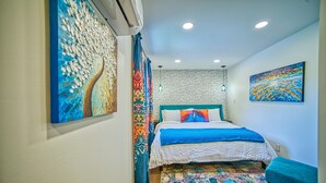 Un dormitorio con cama, cortinas coloridas, alfombra estampada y dos cuadros en las paredes.