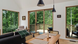 Un moderno salón con una mesa de comedor de madera, sillas negras y beige, y un sofá negro. Amplias ventanas ofrecen vistas a una terraza y árboles.