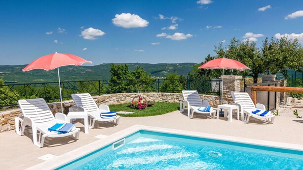 Zona de piscina con sillones blancos y sombrillas rojas, con vistas a un paisaje montañoso.