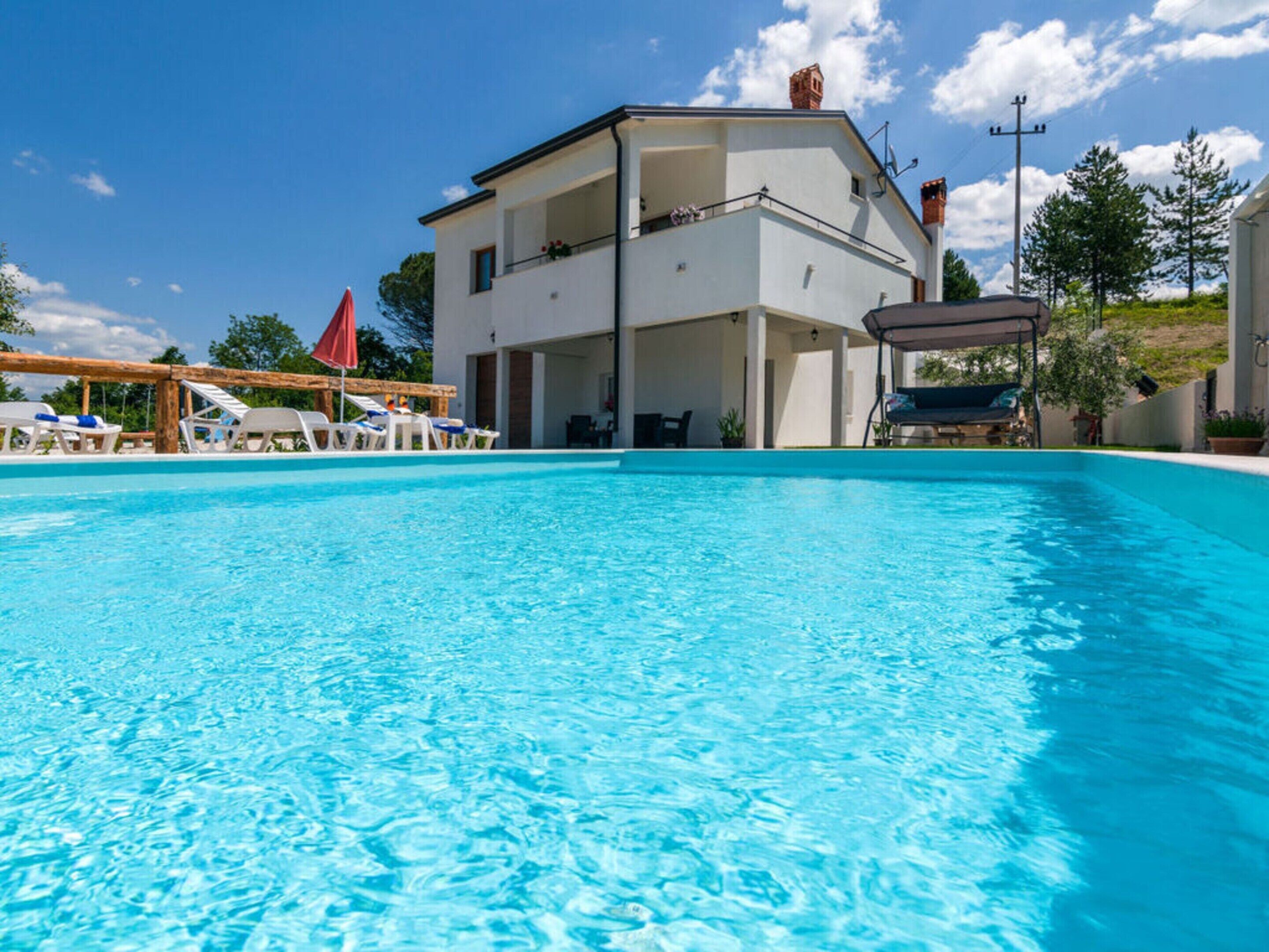 Una piscina con sillas de descanso y sombrillas, un edificio de dos pisos con balcones, y un cielo azul despejado.