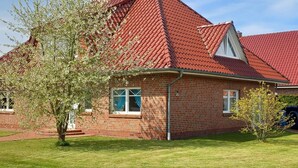 Une maison en briques avec un toit recouvert de tuiles rouges, un petit jardin devant et un arbre au premier plan.