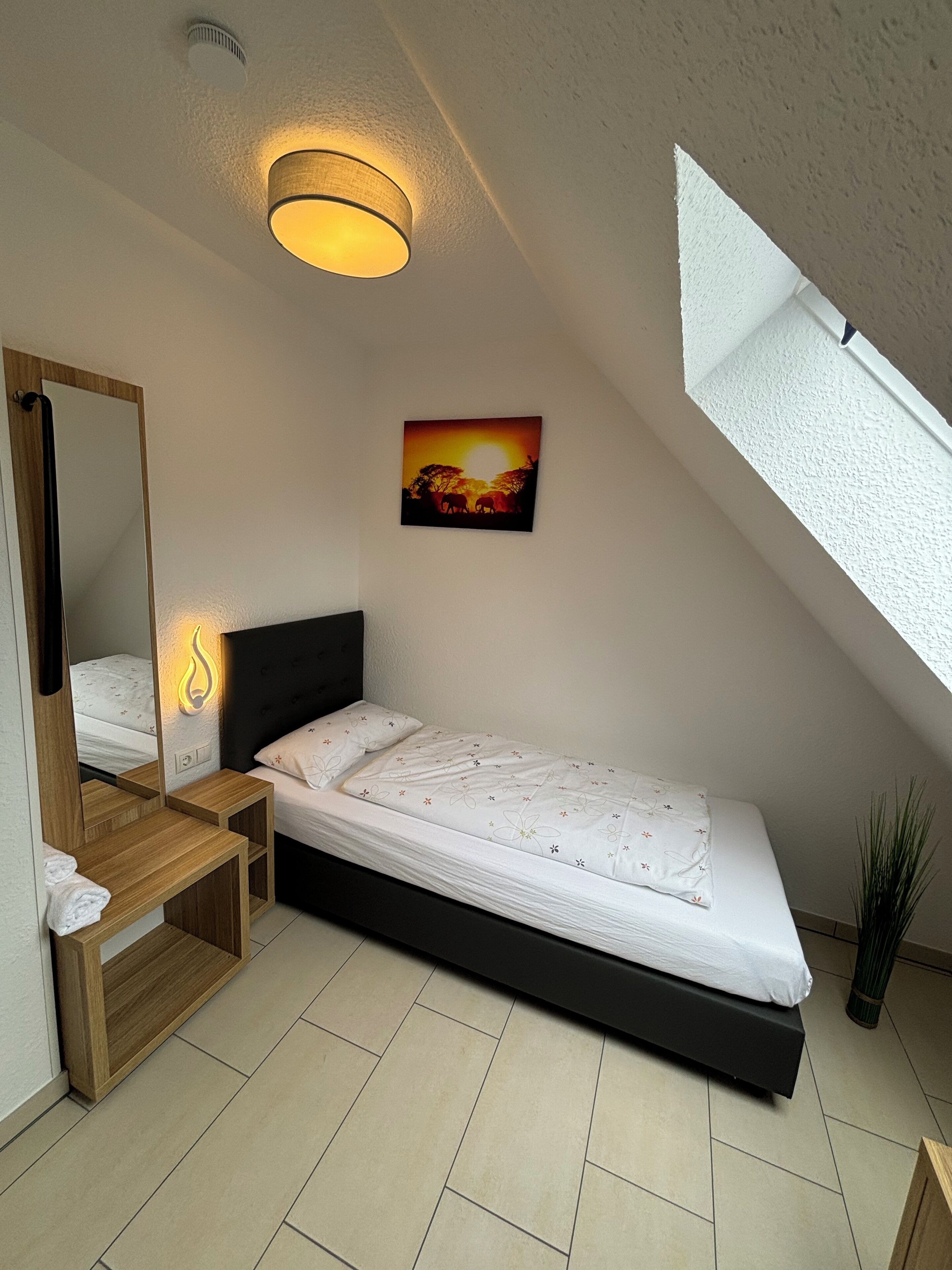 A bedroom with a bed, a bedside table, a mirror, a wall-mounted light, a painting, and a window.