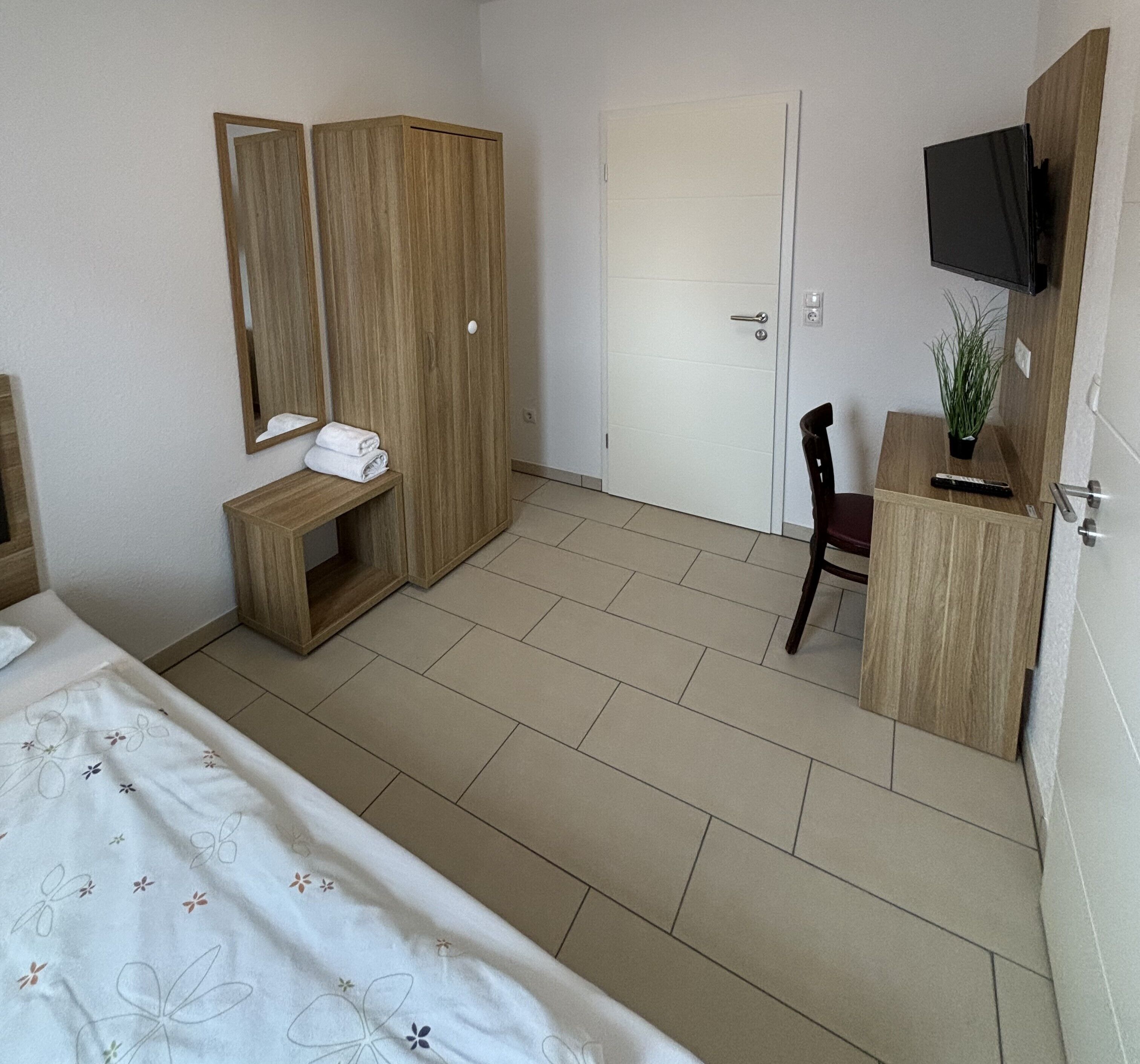 Une chambre d’hôtel avec un lit, une armoire en bois, un bureau avec une chaise, une télévision et une petite plante.