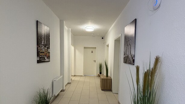 A hallway with tiled flooring, potted plants, and framed pictures on the walls. - Gap Hotel Event & more  (Langwedel)