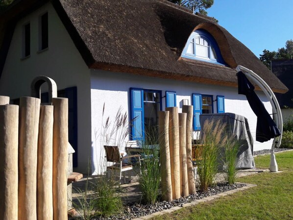Une maison avec un toit de chaume, des murs blancs et des volets bleus. On y trouve une chaise et une table à l’extérieur.