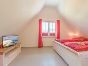 A bedroom with a TV on a wooden shelf, a window with red curtains, and a bed with red bedding. - Adlerhorst Modern Retreat (Glowe)
