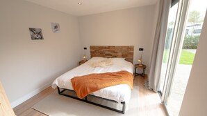 A bedroom with a bed, a headboard made of wood, two bedside lamps, a window with curtains, and a small rug. - Holiday Home in Zevenhuizen With Sauna (Zevenhuizen)