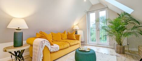 A modern living room with a yellow sofa, two lamps, a green ottoman, and a large window with a view of greenery.