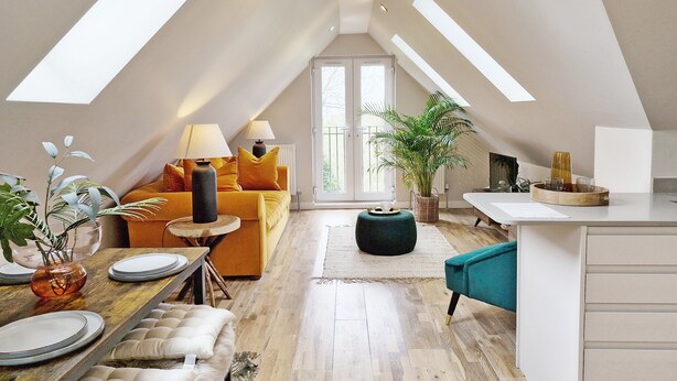 Un loft amplio con techo alto, zona de comedor con mesa y sillas de madera, sofá con cojines, cocina con encimera y un gran ventanal que proporciona luz natural.