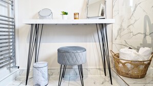 Un baño con tocador blanco, un taburete gris, un cubo de basura de mármol, una cesta de mimbre con toallas y una pequeña planta en maceta.