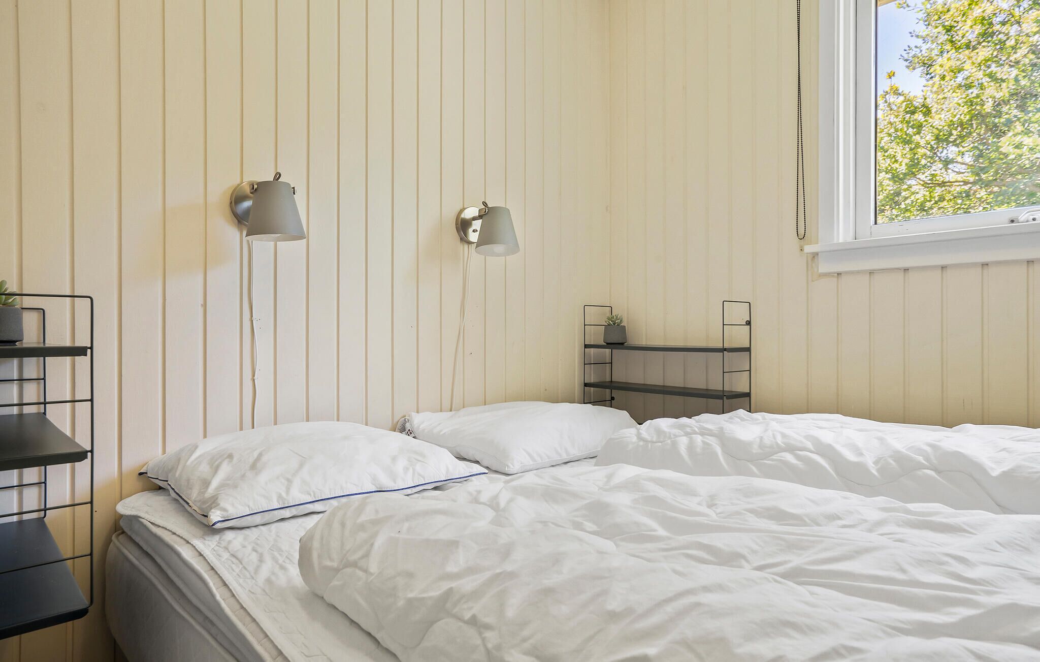 Ein Schlafzimmer mit einem Bett, zwei Wandlampen, einem Fenster mit Blick auf Bäume und einem Regal mit einer Pflanze.