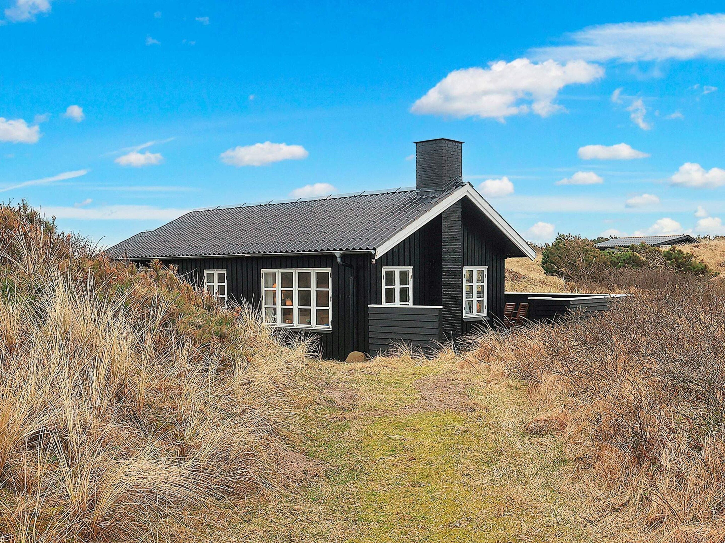 A black wooden house with a sloped roof and a chimney, surrounded by dry grass and shrubs.