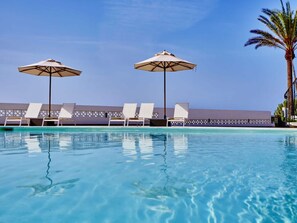 A pool area with lounge chairs and umbrellas, a palm tree, and a building in the background. - Luxurious Ocean Front Holiday Home (San Bartolomé de Tirajana)