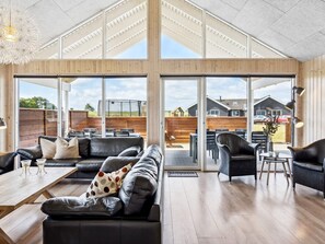 A spacious living area with a large sofa, a coffee table, and a dining set. There are large windows providing a view of the outside.