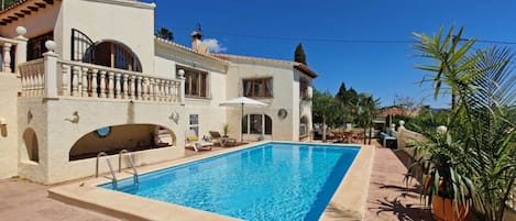 Vue sur une villa de style méditerranéen avec piscine, mobilier d’extérieur et plantes en pot, le tout surplombant la piscine.