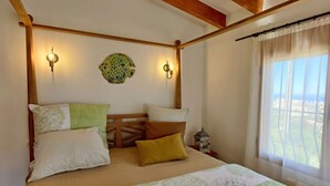 A bedroom with a wooden headboard, a bed with multiple pillows, two wall-mounted lights, a decorative wall piece, and a window with a view. - Villa Fiorida Comfortable Holiday Residence (Calpe)