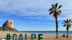 A beach with a large rock formation and palm trees. There is a sign with the hashtag #CALPE. - Villa Fiorida Comfortable Holiday Residence (Calpe)