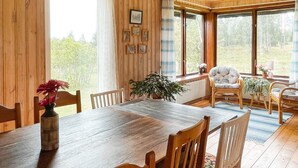 Une table à manger en bois, entourée de chaises, un vase rempli de fleurs et une vue sur l’extérieur grâce à de grandes fenêtres.