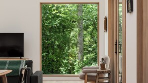 Sala de estar moderna com uma grande porta de vidro deslizante que dá para uma área arborizada, um sofá preto, uma mesa de centro de madeira e uma pequena mesa de madeira com uma planta em vaso.