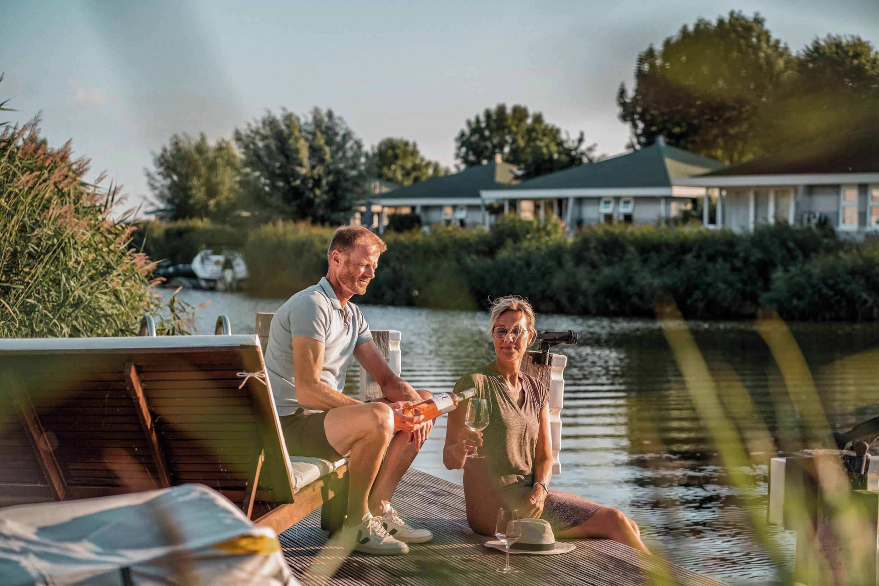 A couple sitting on a dock by a body of water, with a house in the background.