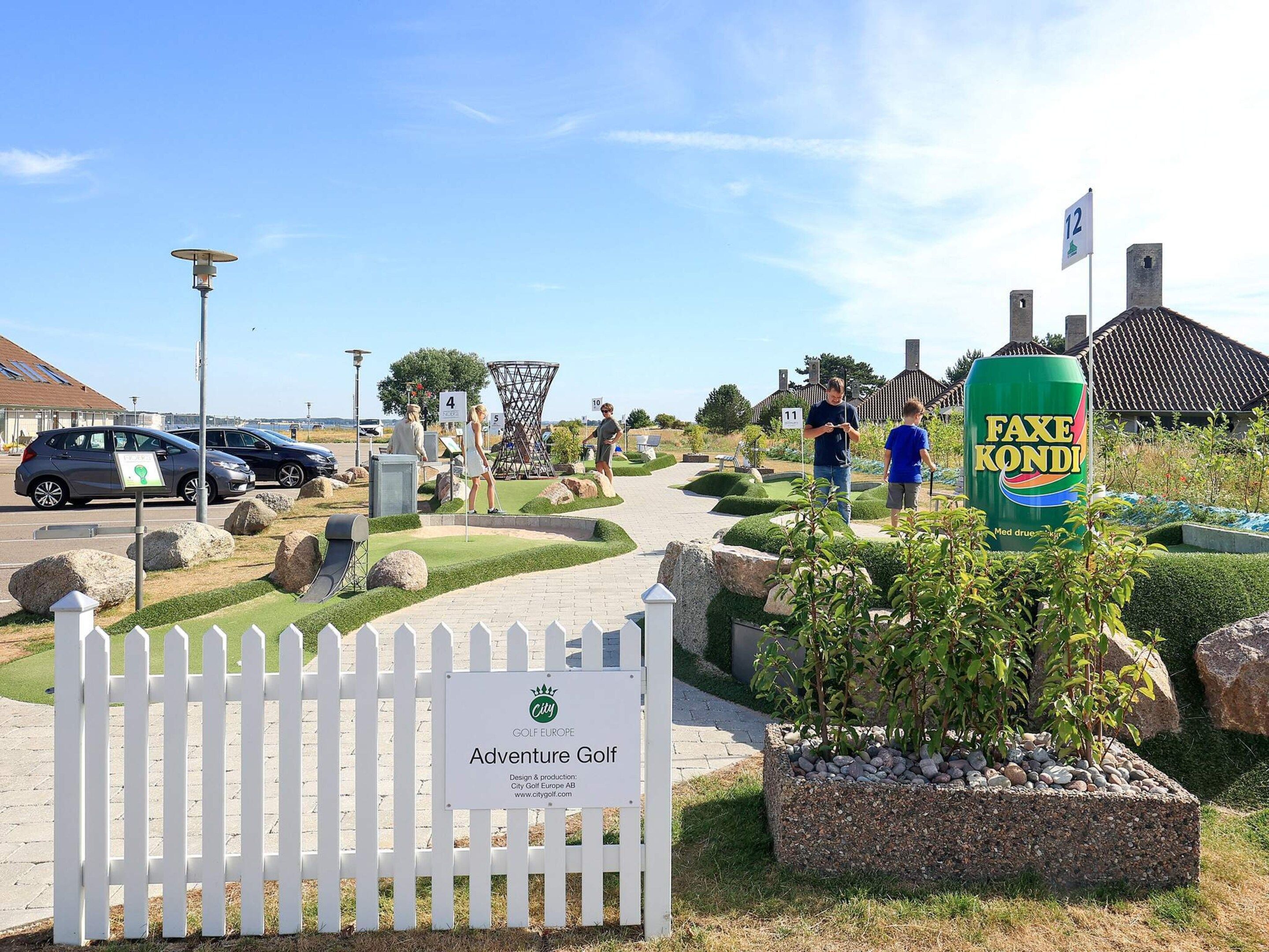 Ingang van een golfbaan met een bordje voor 'Adventure Golf', een witte picket-hek en een blik Faxe Kandi.