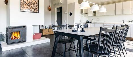 Une cuisine moderne avec des meubles blancs, un coin repas doté d’une grande table et de chaises, et une cheminée où l’on peut voir les flammes.