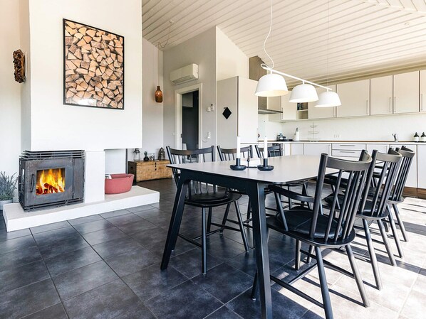 Een moderne keuken met witte kasten, een eethoek met een grote tafel en stoelen, en een open haard met vlammen.