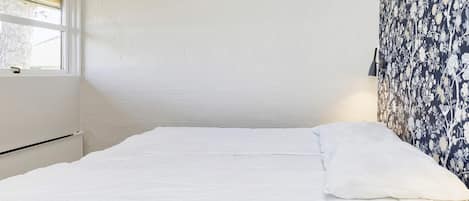 A neatly made bed with white bedding in a room with white walls and a window.