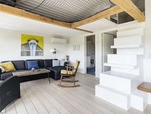 Een moderne woonkamer met een bank, een schommelstoel en een salontafel. Er is een trap met witte treden en een houten leuning.