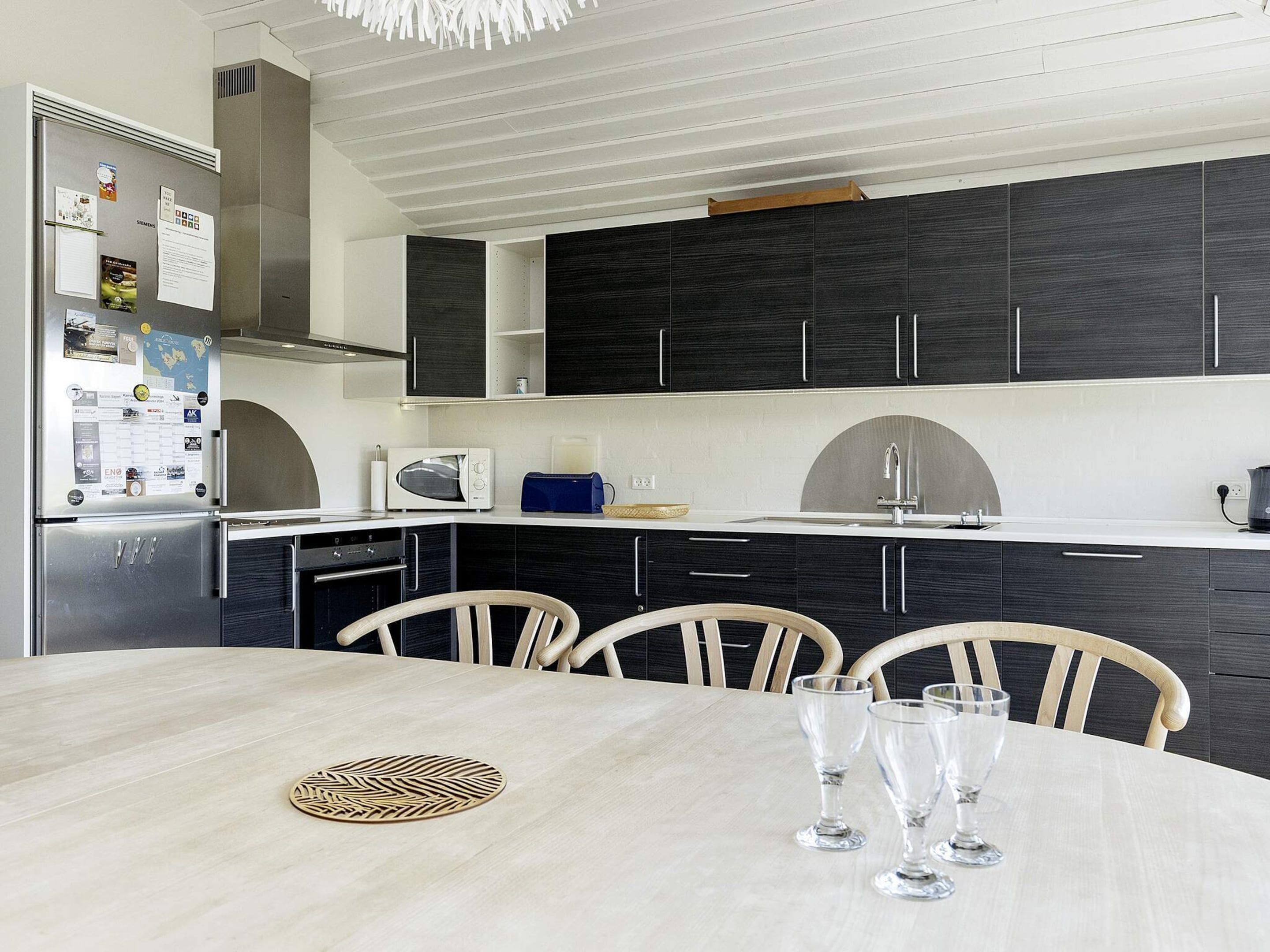 A modern kitchen with dark cabinets, a stainless steel refrigerator, a microwave, and a sink. There is a dining table with chairs and three wine glasses.