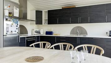 A modern kitchen with dark cabinets, a stainless steel refrigerator, a microwave, and a sink. There is a dining table with chairs and three wine glasses.