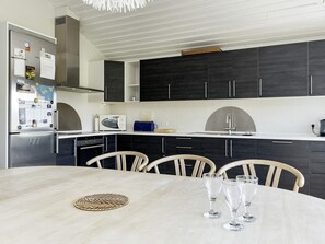 A modern kitchen with dark cabinets, a stainless steel refrigerator, a microwave, and a sink. There is a dining table with chairs and three wine glasses.