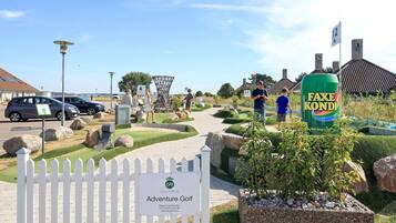 Entrada a un campo de golf con un cartel que dice 'Adventure Golf', una valla de piquetes blancos y una lata de Faxe Kandi.
