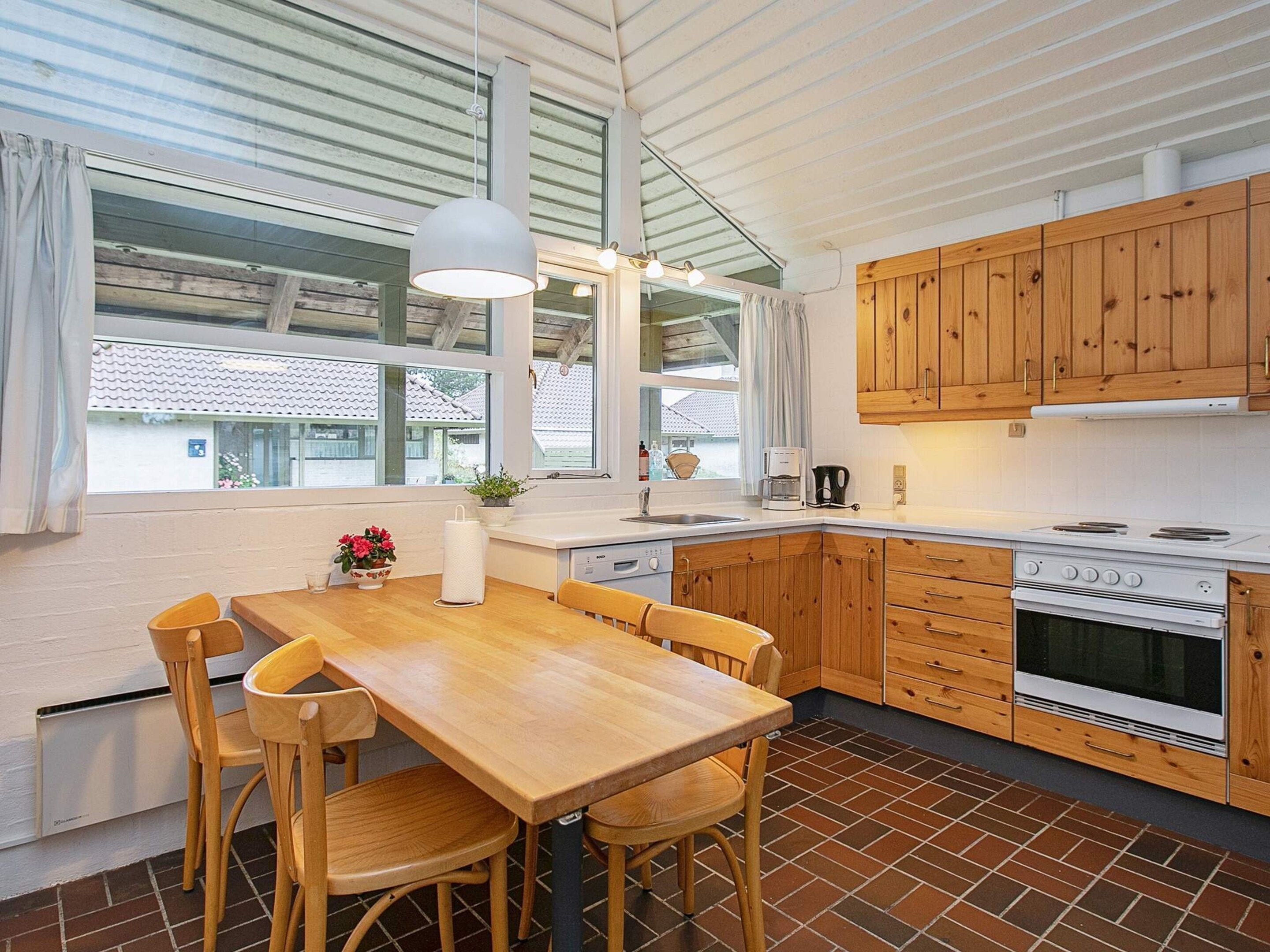 A modern kitchen with wooden cabinets, a large dining table, and a view through the windows.