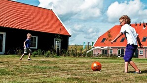 Two children playing soccer on a grassy field with a red-roofed building in the background.