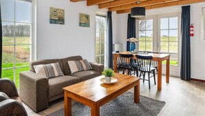 A cozy living room with a sofa, a wooden coffee table, and a dining area with a table and chairs. Large windows offer a view of the outdoors.