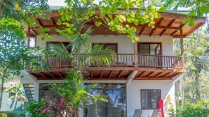 Un bâtiment à deux étages doté d’un balcon en bois, entouré d’une végétation luxuriante et surplombant un ciel dégagé.