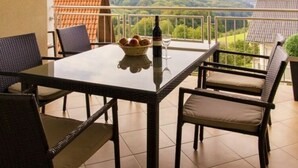 A balcony with a glass table, wicker chairs, a wine bottle, and a bowl of fruit. - Apartment Adlerhorst - Schwalbenhof Farm (Sankt Stefan im Rosental)