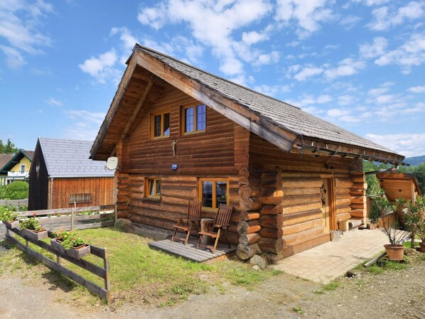 A wooden log cabin with a sloped roof, a porch with chairs, and a number '196' on the front wall.