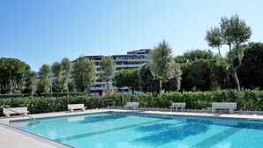 Una piscina con sillones de descanso y un banco, rodeada de follaje y un edificio de varios pisos al fondo.