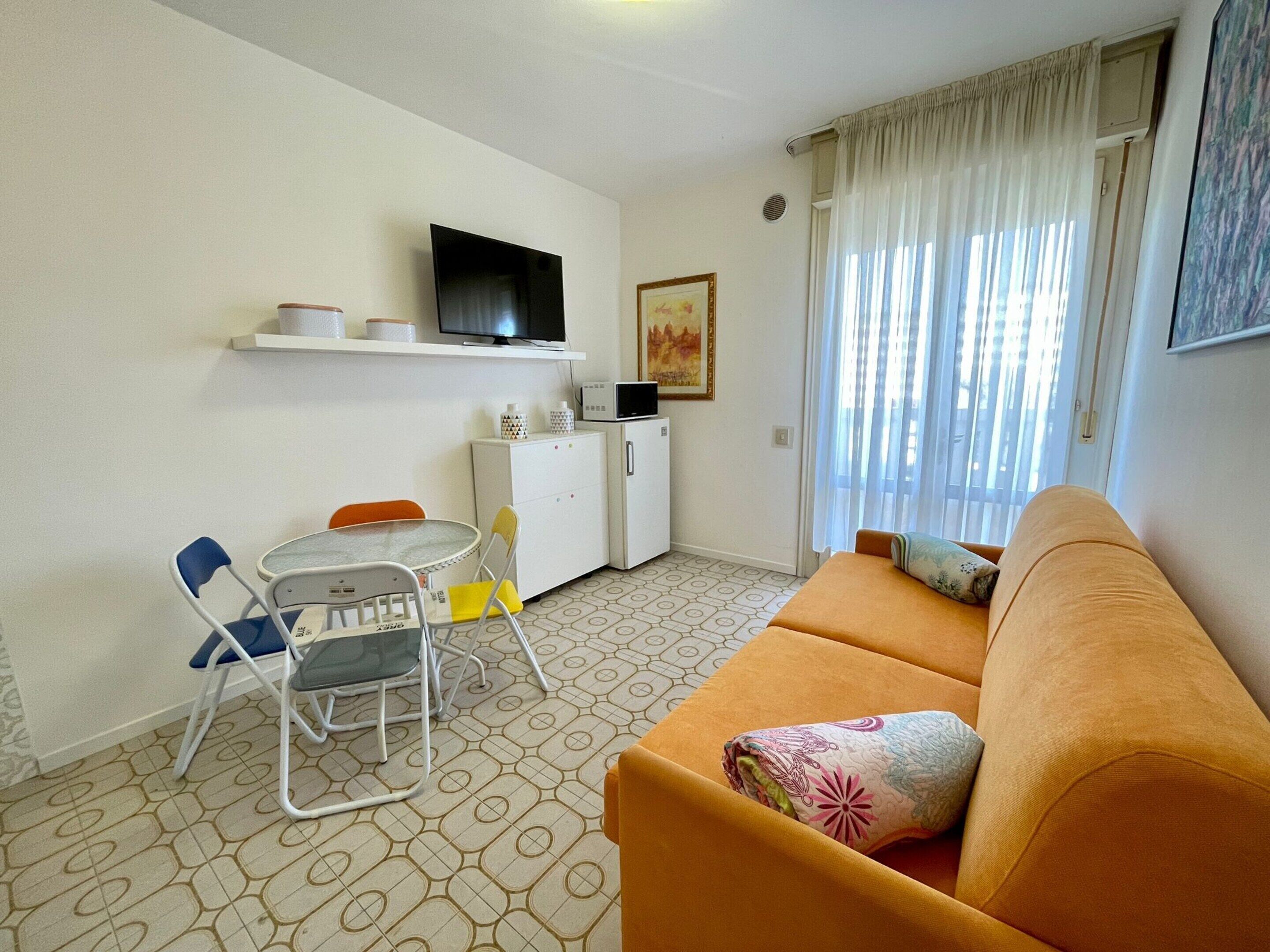 A compact living space with a sofa, a dining table with chairs, a TV mounted on the wall, a small refrigerator, and a window with curtains.