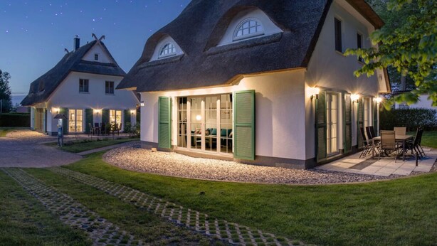 A traditional house with a thatched roof, large windows, and a well-lit outdoor area. - Droom Unter Reet 2 Comfortable Holiday Residence (Rerik)