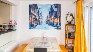 A dining area with a table covered by a map-themed tablecloth, a painting of a snowy street on the wall, and a yellow curtain on the window. - Duplex Apartment Near the Center (Middelkerke)