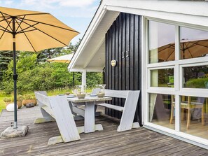 Um deck de madeira com uma mesa, cadeiras, um guarda-sol grande e uma casa com revestimento preto e janelas amplas.