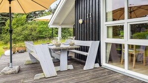 Une terrasse en bois avec une table et des chaises, un grand parasol, et un bâtiment à l’extérieur noir et aux ornements blancs.