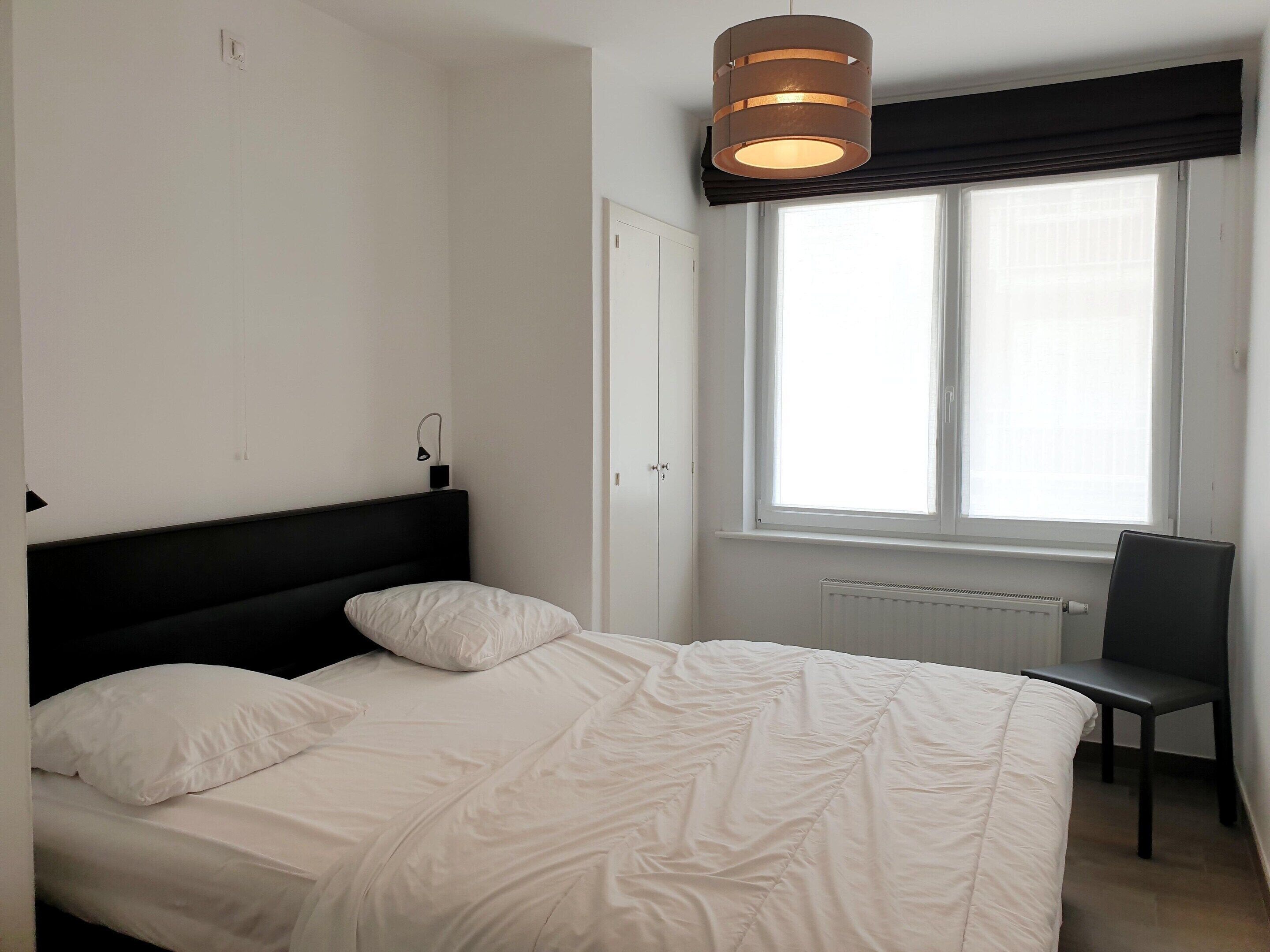 A bedroom with a bed, a chair, a window with blinds, and a wall-mounted light fixture.