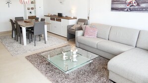 A modern living room with a grey sofa, a glass coffee table, and a dining area with a wooden table and chairs. - Apartment in Nieuwpoort by the Beach (Nieuwpoort)