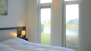 A bedroom with a bed, a nightstand, a lamp, and a window with a view of a building and greenery. - Chalet in Zeeland by the Lake (Wemeldinge)