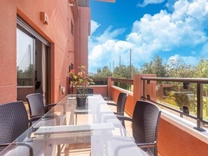 Ein Balkon mit Glastisch und Stühlen, einem Topfpflanzen und Blick auf Grünflächen und einen klaren Himmel.